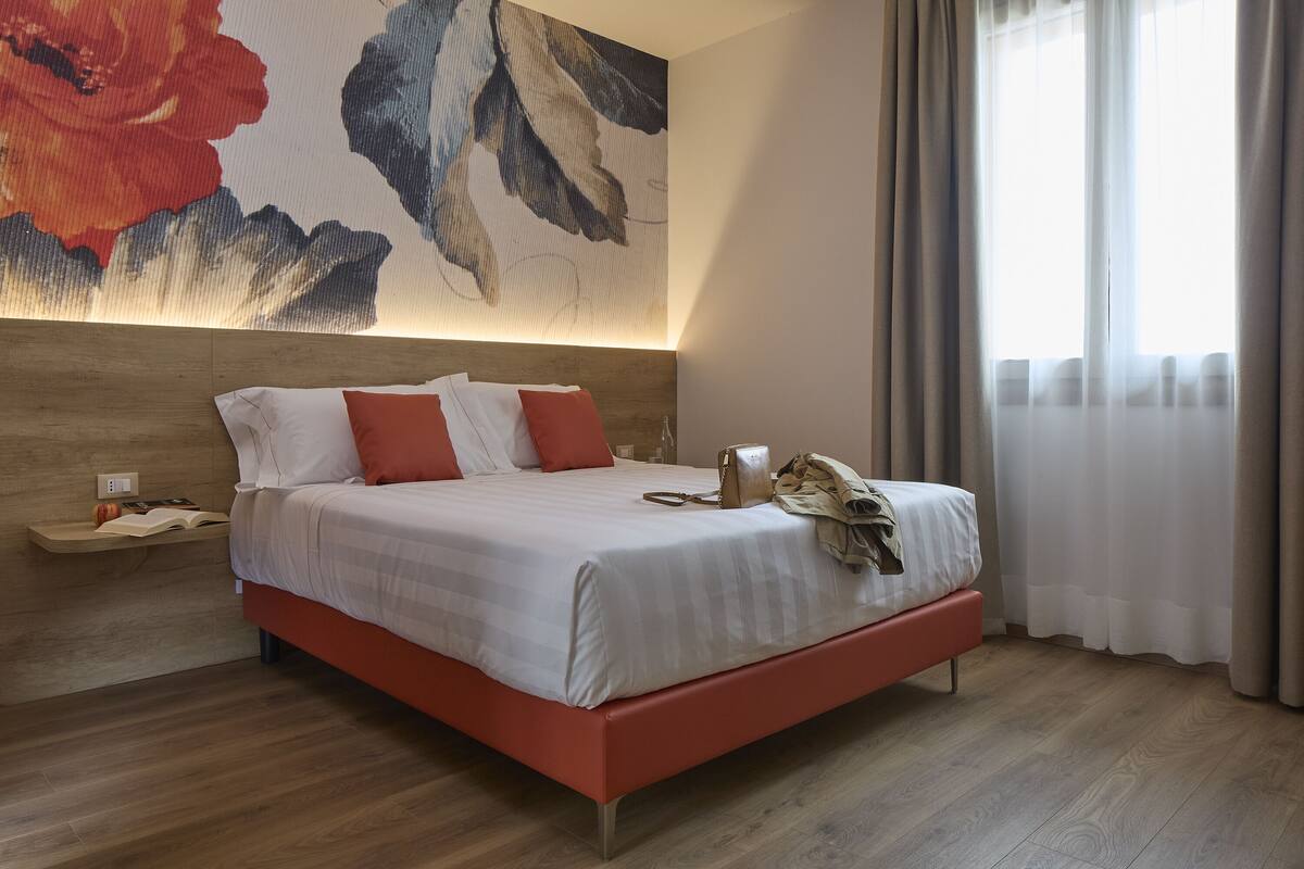 A modern hotel room with a large bed, a wooden headboard, a nightstand, and a large floral artwork on the wall.