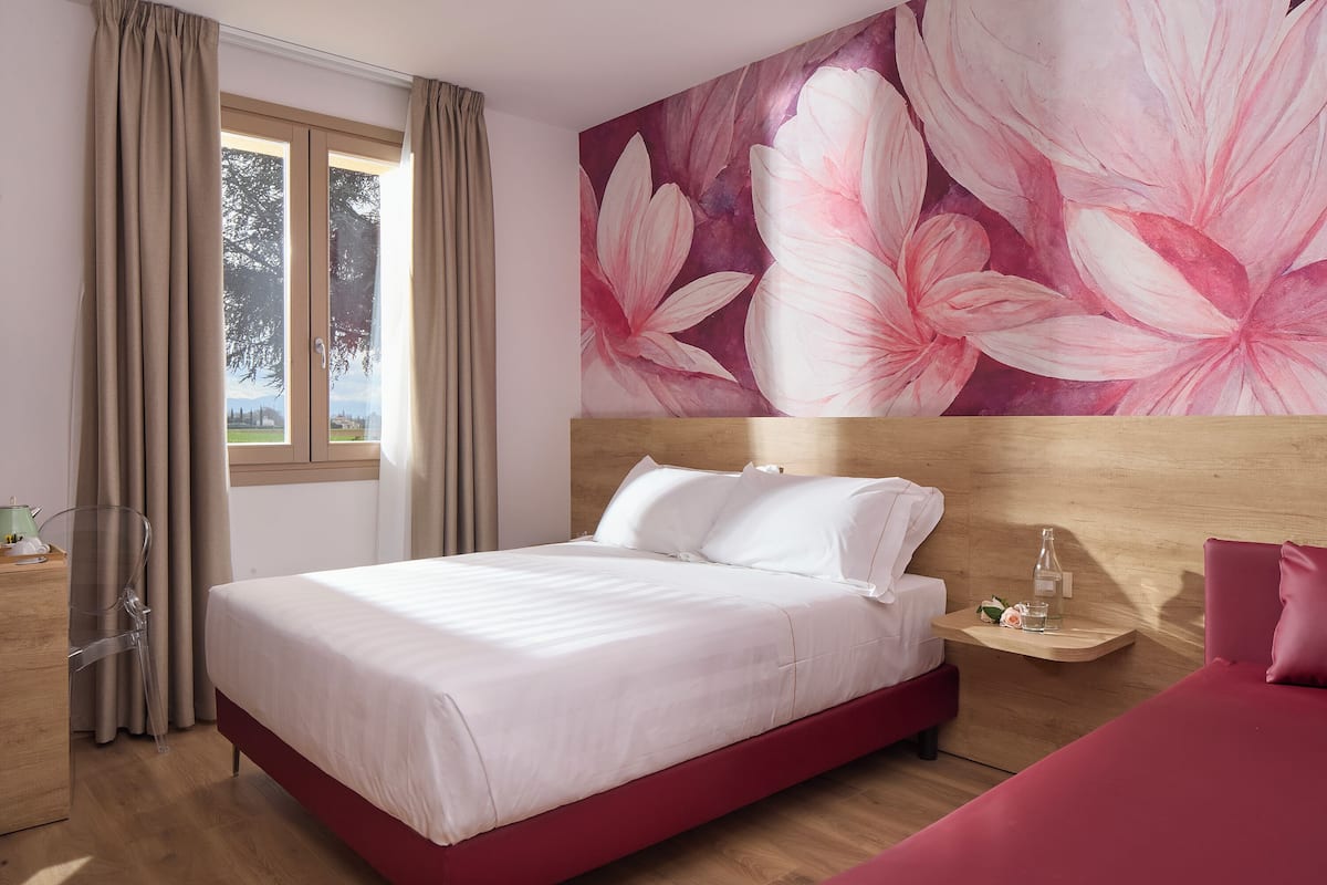 A modern hotel room with a large bed, a wooden headboard, a bedside table with a bottle and glass, and a view of the outdoors through a window with curtains.