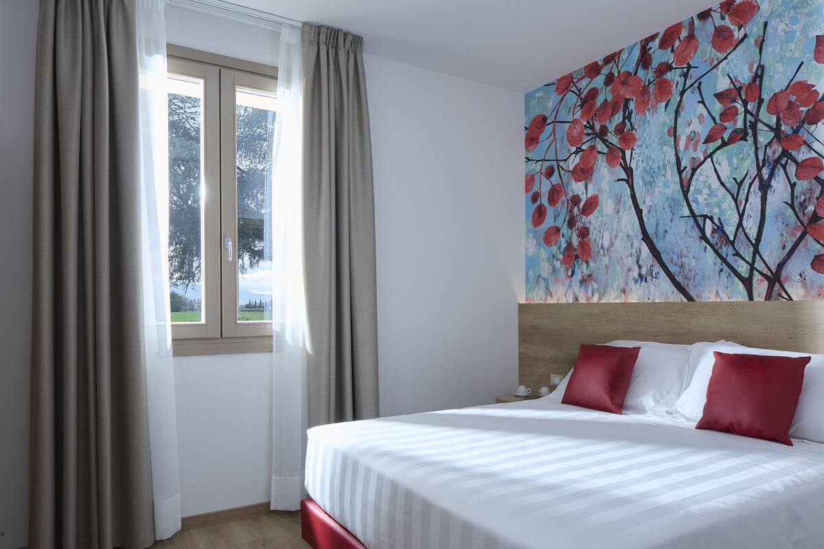 A hotel room with a large bed, beige curtains, a window with a view of trees, and a painting of red leaves on the wall.
