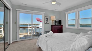 A bedroom with a bed, a ceiling fan, a nightstand, a lamp, and a view of the ocean through large windows. - Waterfront Coastal Retreat w/ Pool | Sunset Views! (North Topsail Beach)