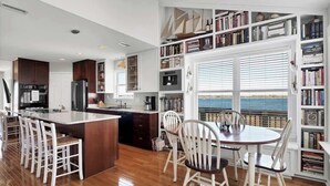 A modern kitchen with a central island, white bar stools, and a dining table with chairs. There is a large window with blinds, a bookshelf filled with books, and a view of the sea. - Waterfront Coastal Retreat w/ Pool | Sunset Views! (North Topsail Beach)