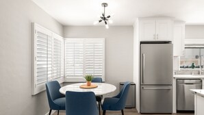 A modern kitchen with white cabinets, a stainless steel refrigerator, and a dining area with blue chairs and a round table. - Ocean Views w/ Private Ground Floor Patio | Fire Pit (San Diego)