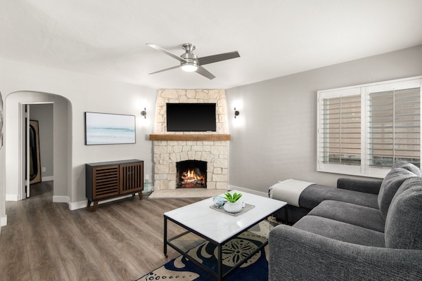 Stylish living room with a stone fireplace, wooden mantel, and mounted TV. Modern gray sectional sits beside a marble coffee table. Ceiling fan enhances comfort and airflow.