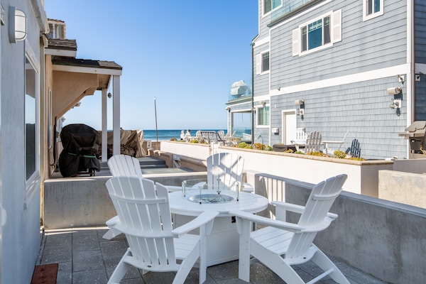 Coastal patio with white Adirondack chairs around a modern fire pit table. Ocean views, a built-in grill, and neighboring beach homes enhance the setting. Easy beach access.
