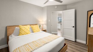 A bedroom with a bed, a ceiling fan, a nightstand with a lamp, and a door leading to a kitchen. - Ocean Views w/ Private Ground Floor Patio | Fire Pit (San Diego)