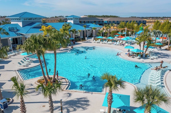 A large outdoor swimming pool surrounded by palm trees and lounge chairs, with a view of hotel buildings in the background.