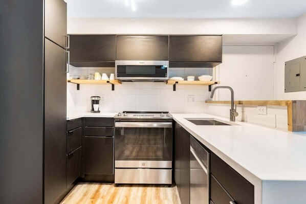 Una cocina moderna con electrodomésticos de acero inoxidable, microondas, horno y lavavajillas. Cuenta con una encimera de madera y un lavamanos con grifo.