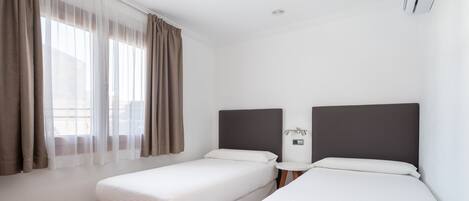 A hotel room with two single beds, a small bedside table, a wall-mounted air conditioner, and a window with sheer curtains.