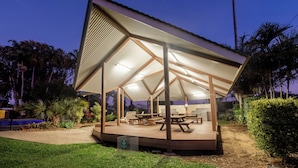 A covered outdoor area with picnic tables and benches, surrounded by greenery and palm trees.