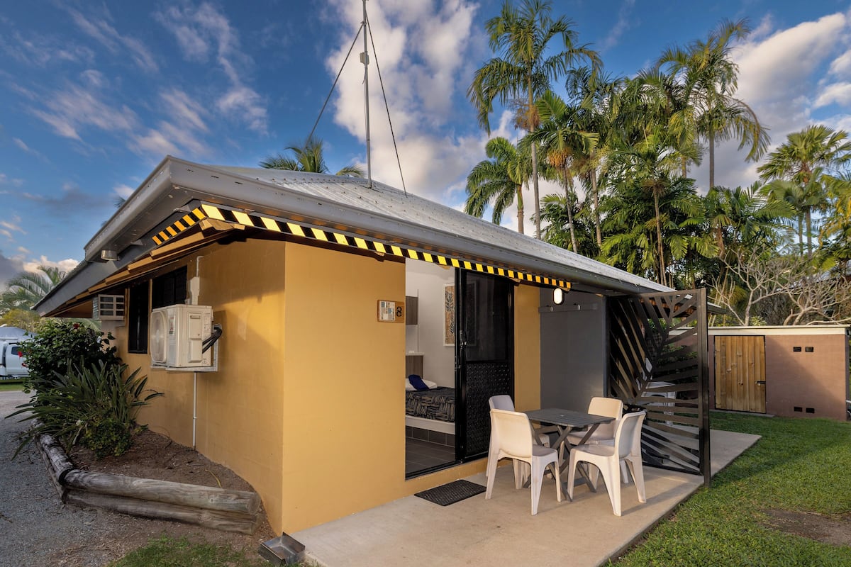 A small, modern outdoor structure with a table and chairs, surrounded by palm trees and a clear sky.