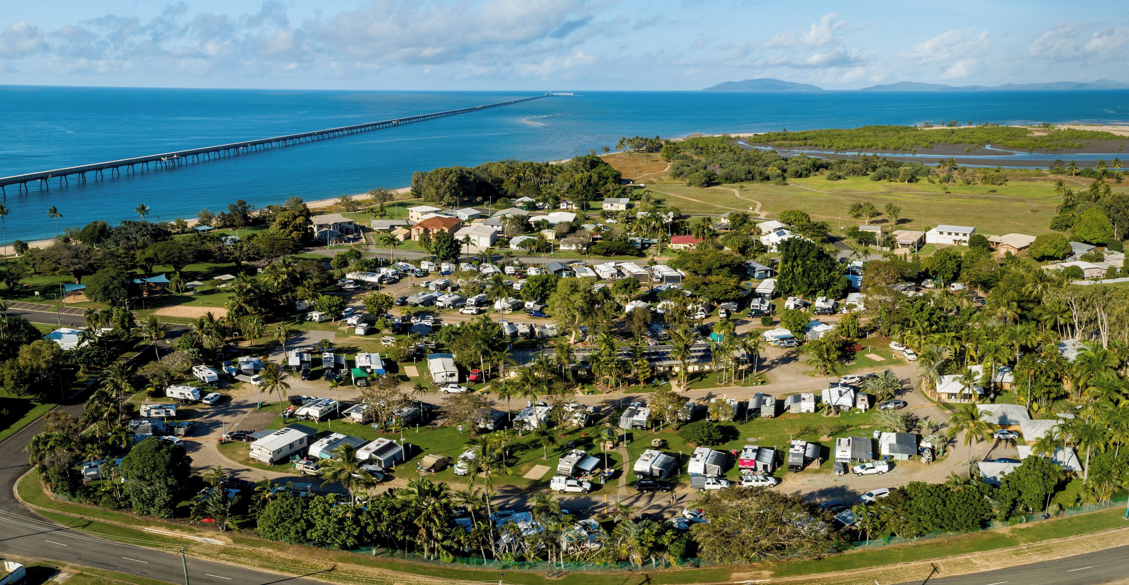 An aerial view of a campground with RVs and trailers, a bridge in the background, and a beach with palm trees.