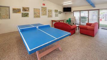 A ping pong table, two red chairs, a small table, and wall-mounted fish illustrations in a room with a speckled floor and a television on the wall.