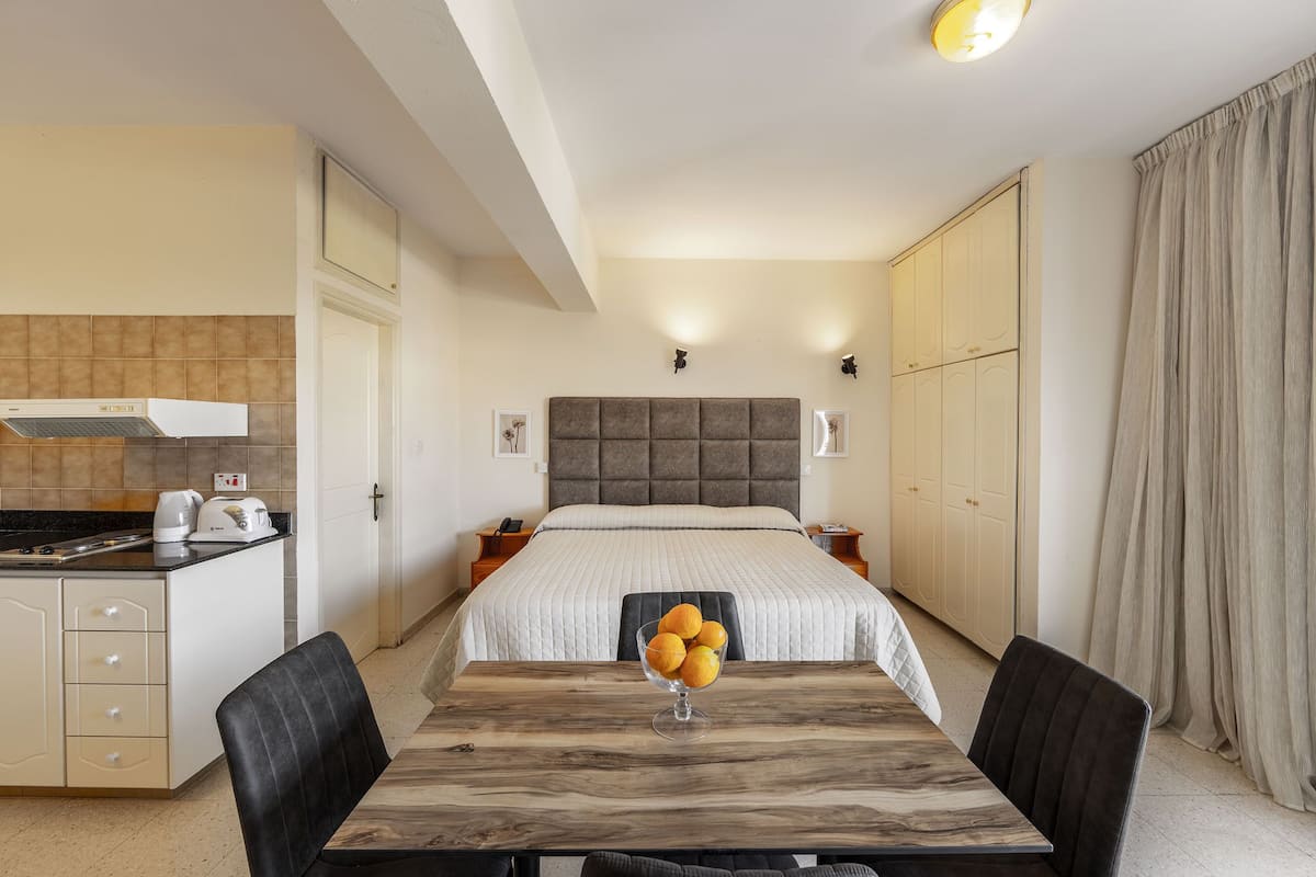 A modern hotel room with a large bed, a wooden dining table with chairs, a kitchen area, and a wardrobe.