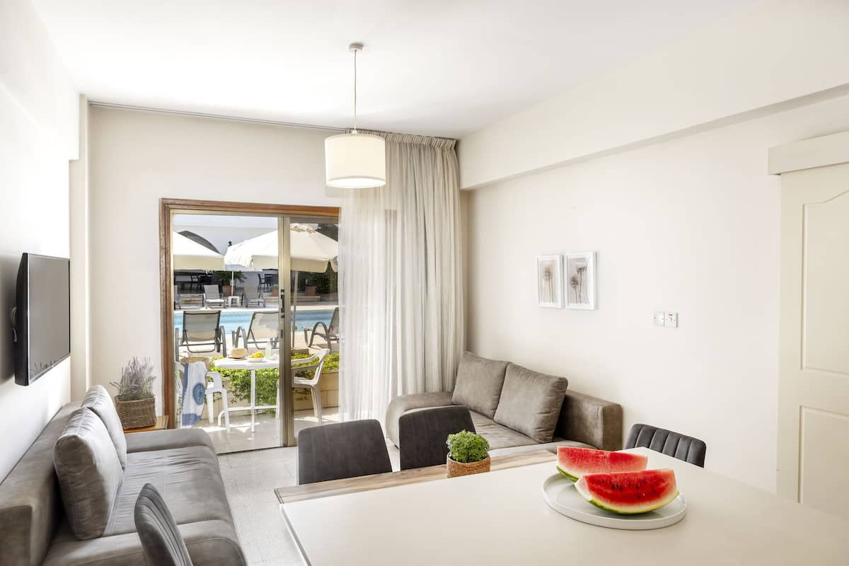 A modern living room with a sofa, dining table, and chairs. There is a large window with a view of an outdoor pool area.