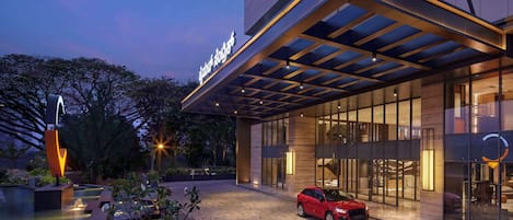 A modern hotel with a covered entrance, a swimming pool, and a red car parked in front.
