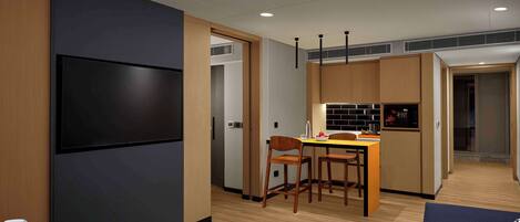 Una habitación de hotel moderna con un televisor de pantalla plana grande, un área de comedor con una pequeña cocina y un rincón de descanso acogedor con un sofá y una mesa.