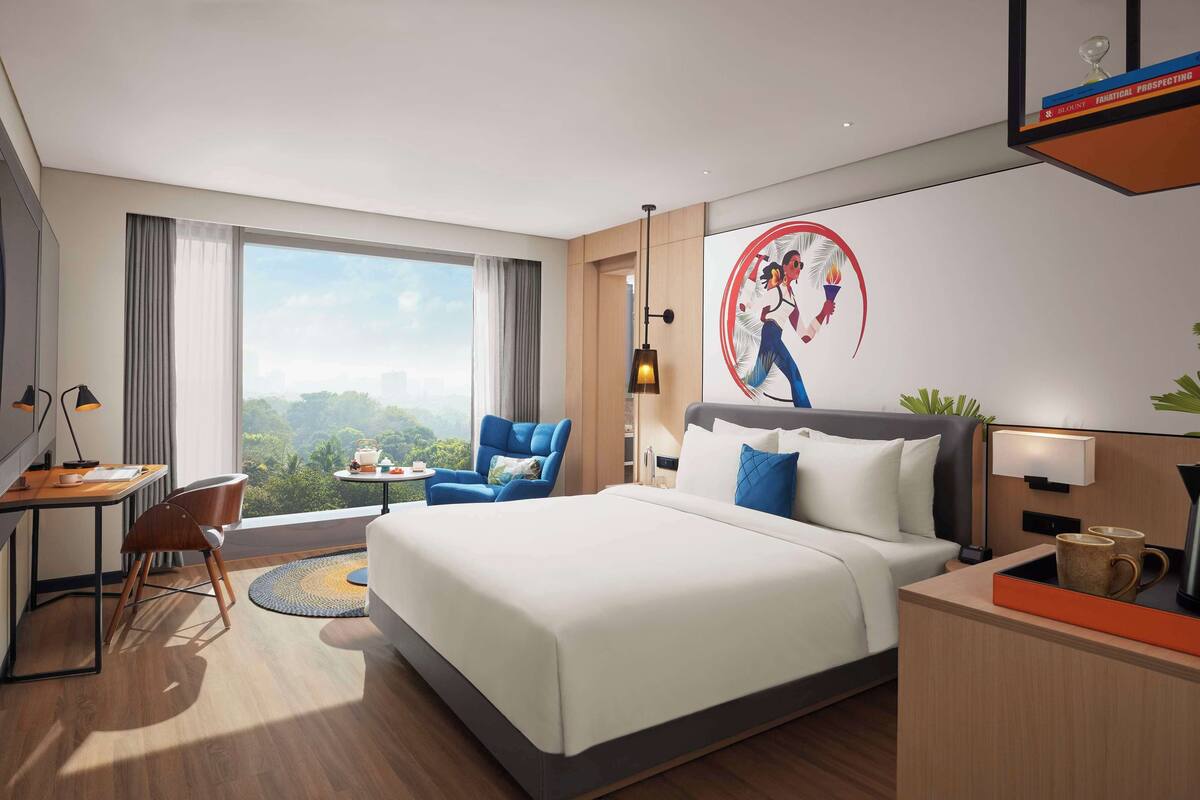 Une chambre d’hôtel moderne avec un grand lit, un bureau avec une chaise, un fauteuil bleu et une vue sur la ville.