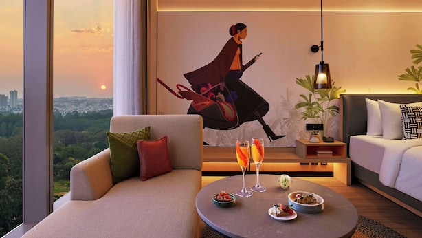 A modern living room with a large window, a sofa, a round coffee table with drinks and snacks, a painting on the wall, and a side table with a lamp. - Hyatt Centric Hebbal Bengaluru (Bengaluru)