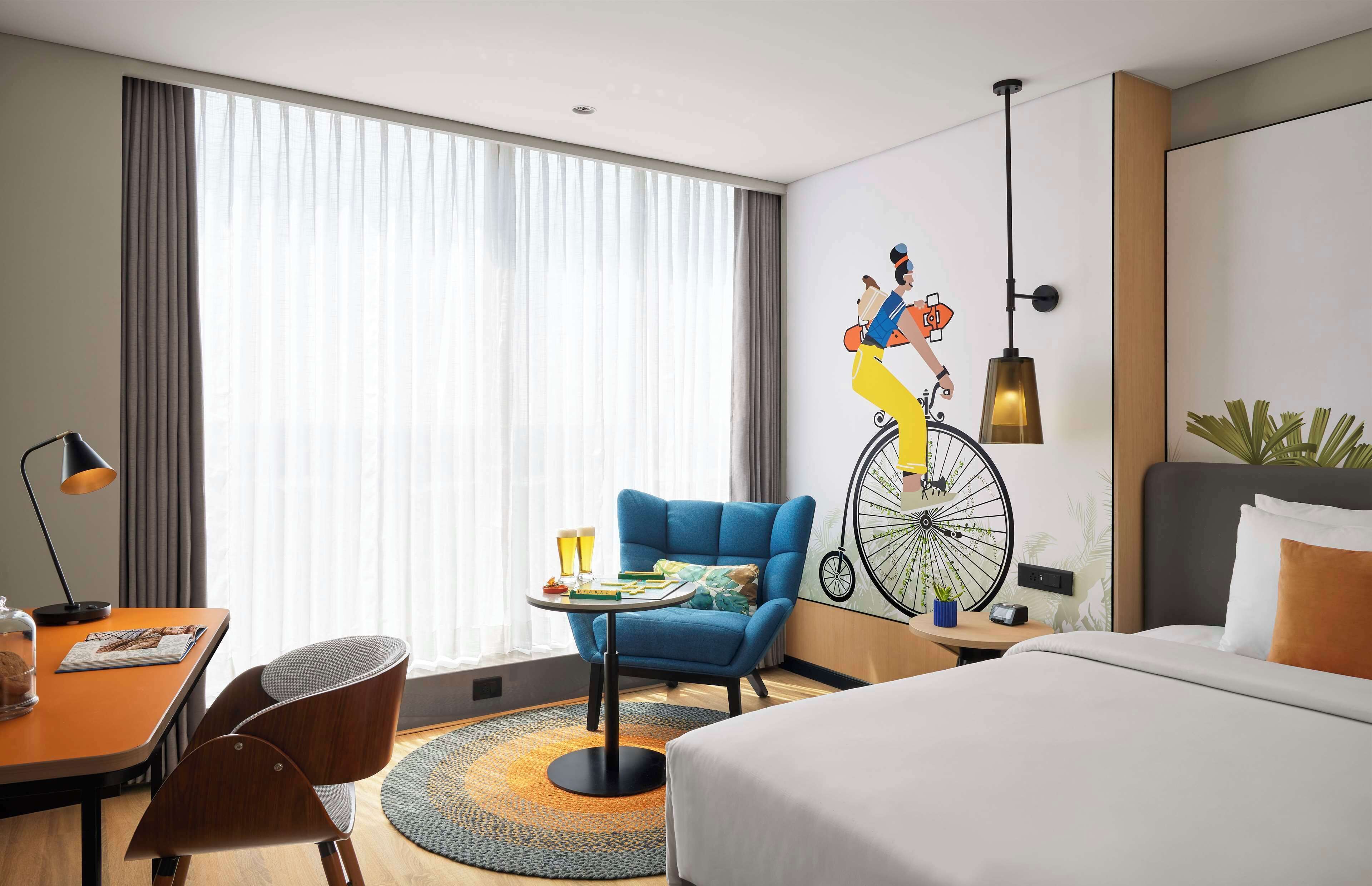 A hotel room with a bed, a desk with a lamp, a chair, a small table with drinks, and a wall mural.