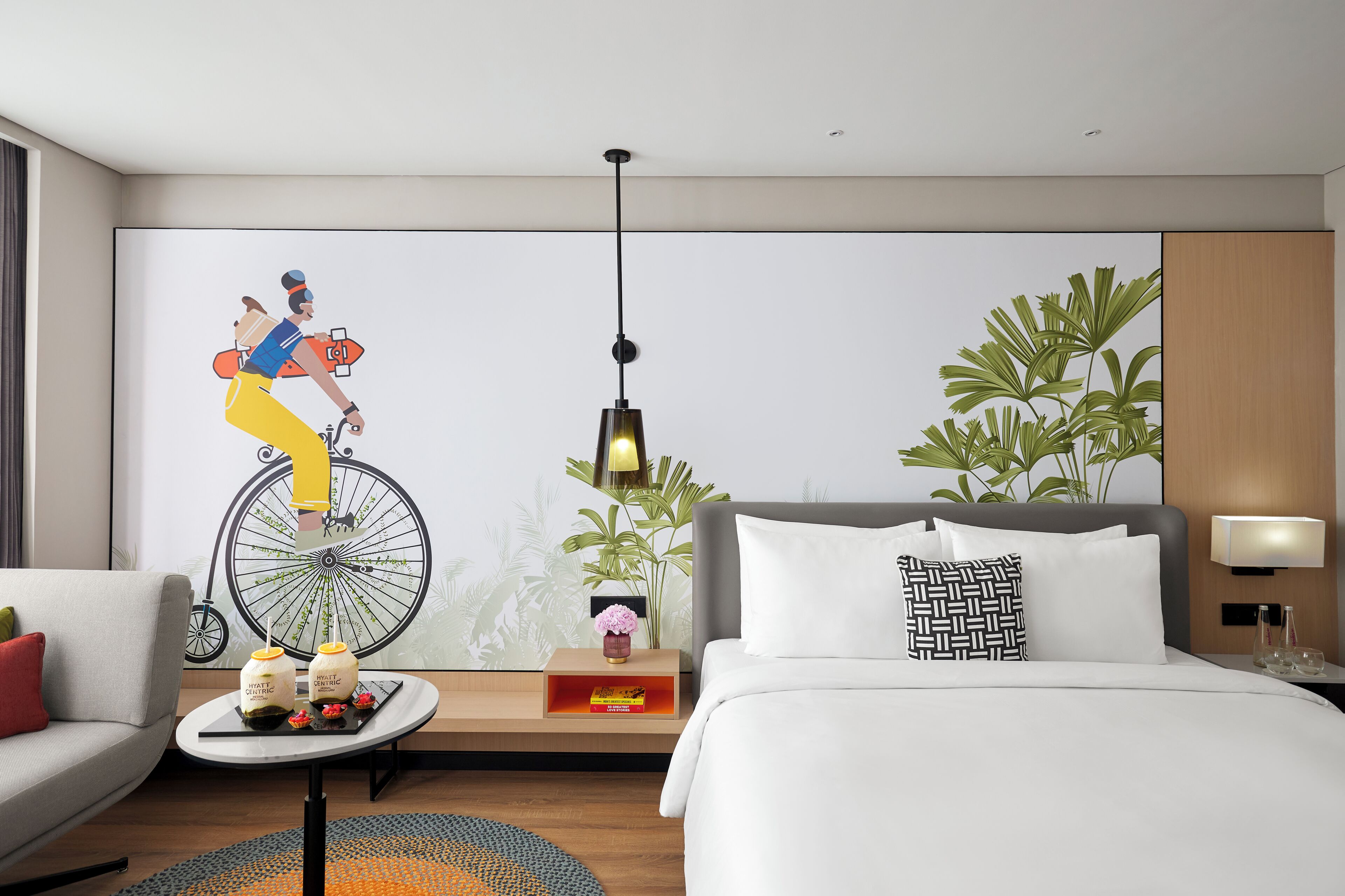 Une chambre d’hôtel moderne avec un grand lit, un canapé, une petite table agrémentée d’objets décoratifs, une fresque murale et une plante en pot.