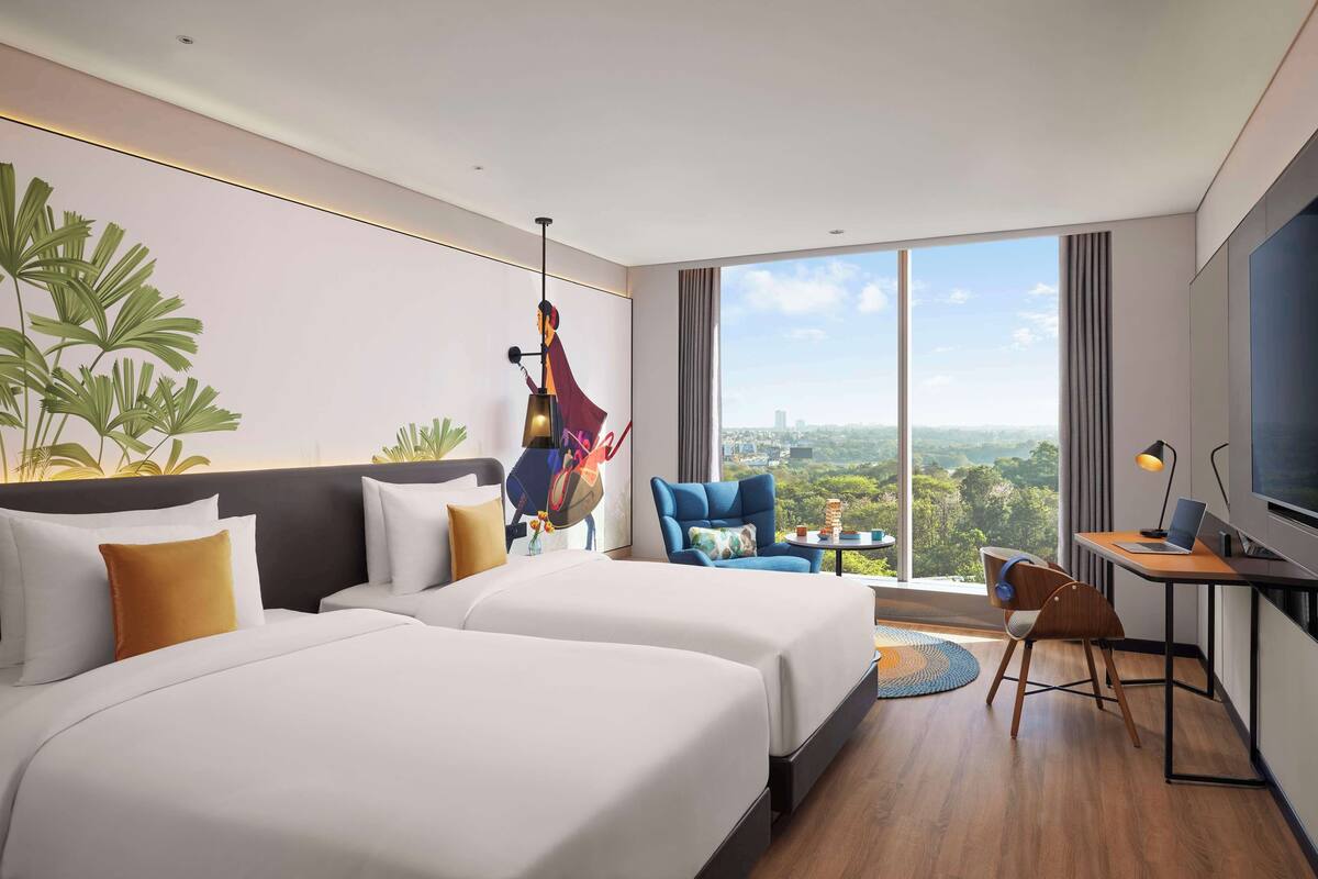 Une chambre d’hôtel avec deux lits, un bureau avec un ordinateur portable, une télévision et une grande fenêtre donnant sur la ville.