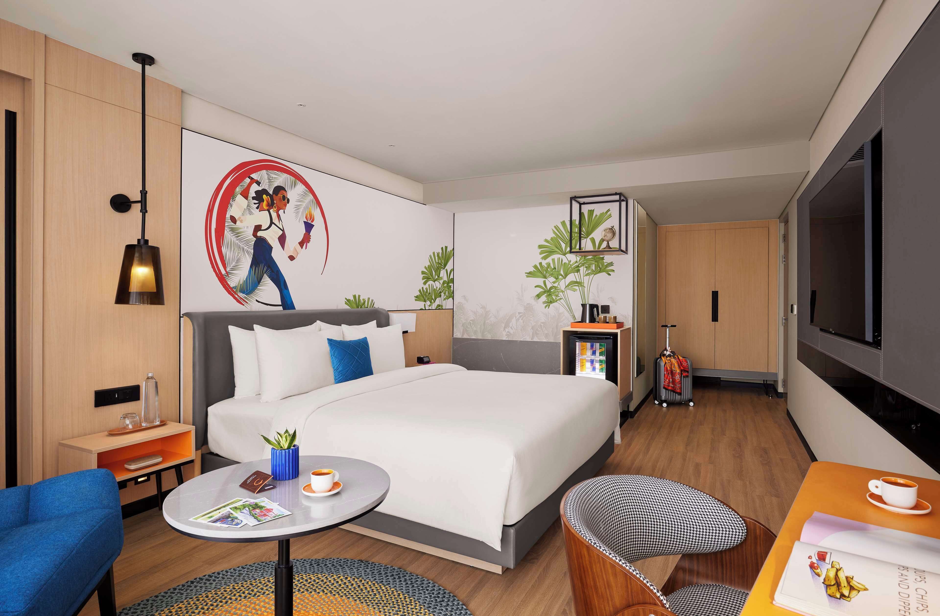 A modern hotel room with a large bed, a desk with a chair, a small table with a cup, a TV, and a suitcase.