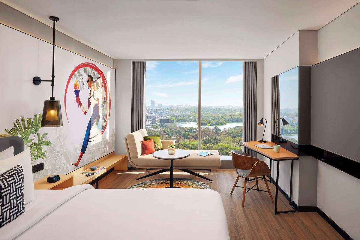Une chambre d’hôtel moderne dotée d’un grand lit, d’un bureau avec une chaise, d’un canapé, d’une télévision et d’une grande fenêtre donnant sur la ville.