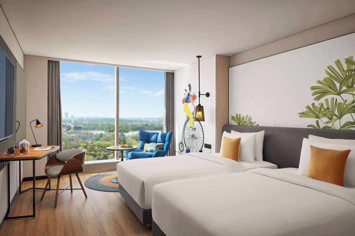 Une chambre d’hôtel avec deux lits, un bureau avec une chaise, une grande fenêtre donnant sur la ville et une décoration murale représentant un vélo.