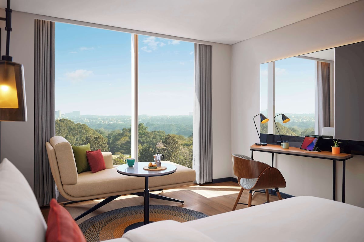 Une chambre d’hôtel moderne dotée d’une grande fenêtre donnant sur la ville, d’un canapé, d’une petite table ronde, d’un bureau avec un ordinateur portable et d’une chaise.