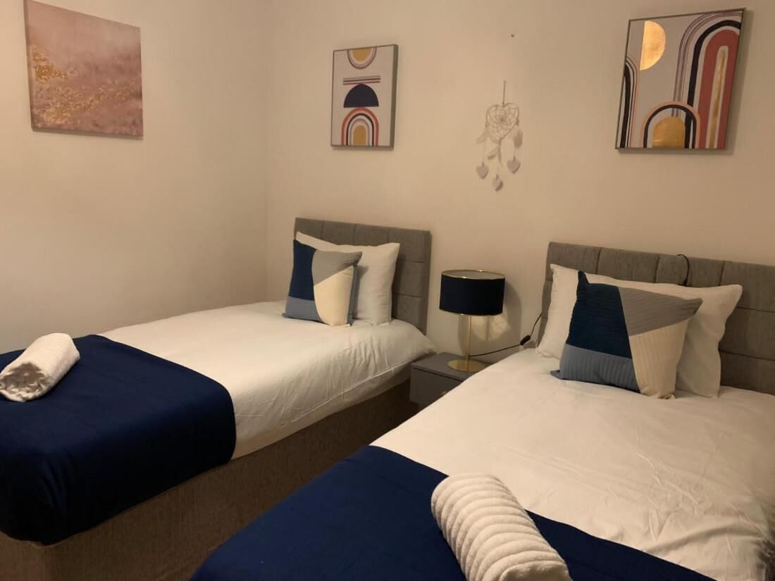 Una camera d'albergo con due letti, ciascuno con copripiumino bianco e blu, una lampada da comodino e un tavolino tra i letti.