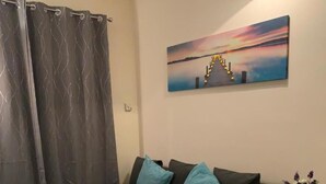 Uma sala de estar moderna com um sofá escuro, almofadas azul-turquesa, uma mesa de centro de vidro e uma pintura de um pôr do sol.