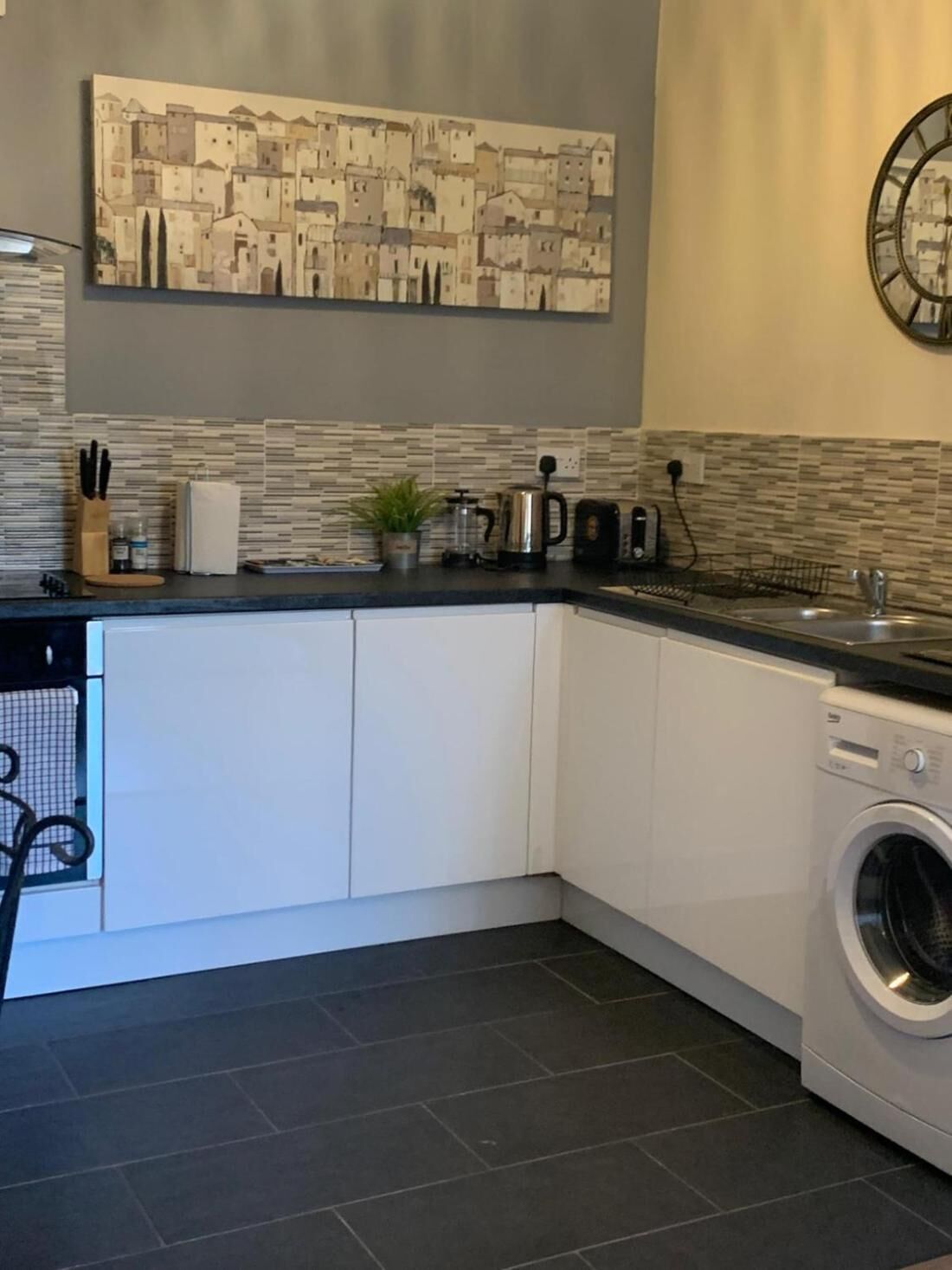 A compact kitchen with a built-in washing machine, a tiled backsplash, and a wall-mounted clock.