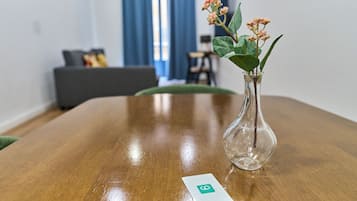 A wooden table with a clear glass vase containing pink flowers, a business card, and a blurred background of a room with a sofa and a window.