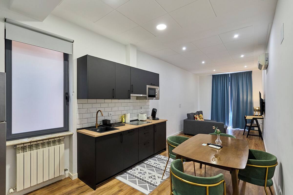 Une cuisine moderne avec des meubles de couleur sombre, un four à micro-ondes et un évier. On y trouve également un coin repas avec une table en bois et des chaises vertes.