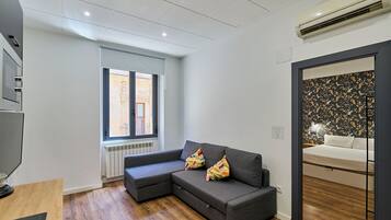 Una sala de estar moderna con un sofá seccional gris, piso de madera, un ventanal con vista a un edificio y un equipo de aire acondicionado en la pared.