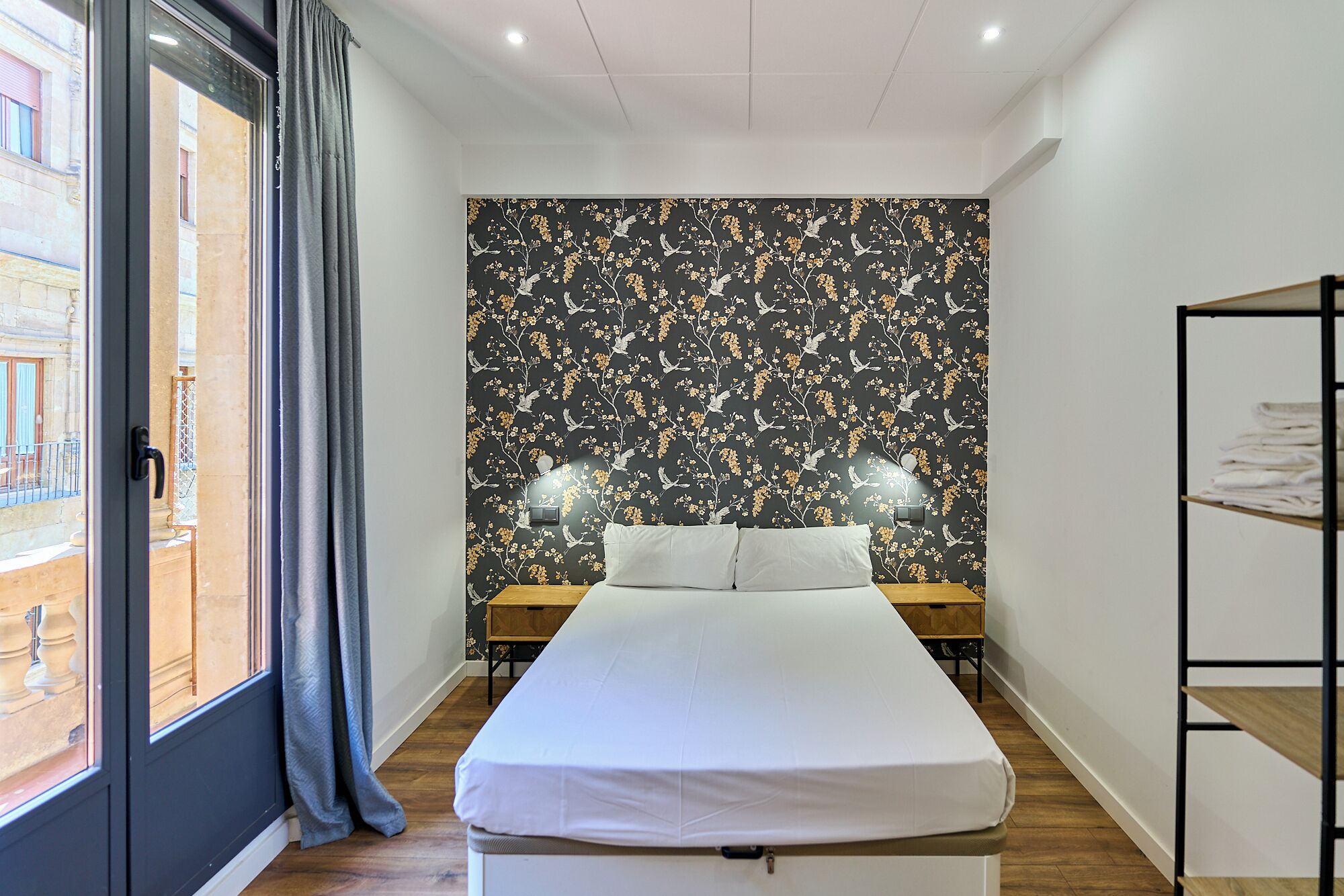 Una habitación de hotel moderna con una cama grande, una mesita de noche de madera, un armario y una ventana con vistas al exterior.