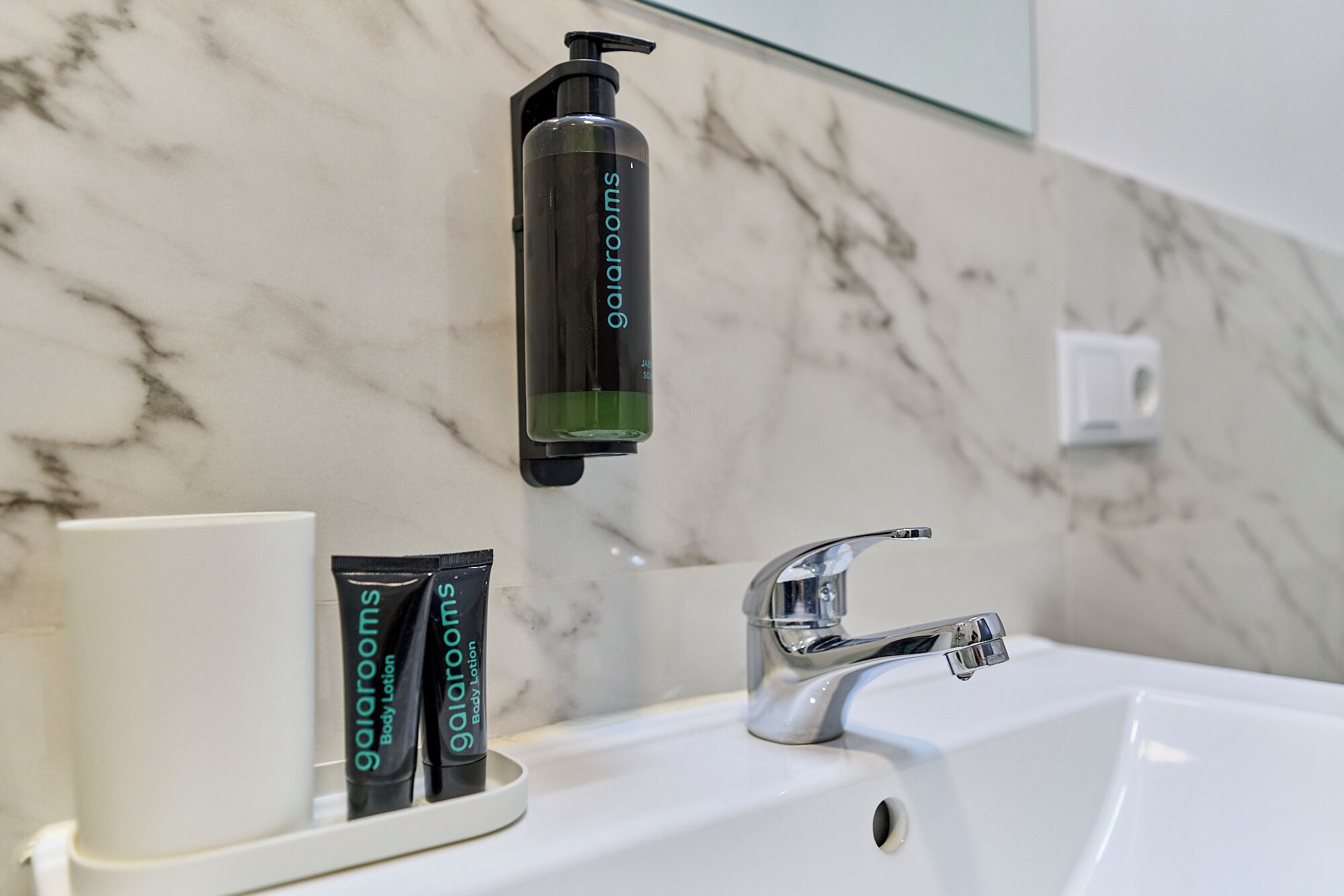 A bathroom with a marble wall, a white sink, a chrome faucet, and two tubes of body lotion labeled 'gairoom'.