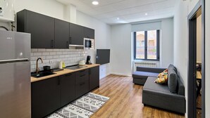 A modern kitchen with dark cabinets, a stainless steel refrigerator, a microwave, and a wooden countertop. There is a gray sofa with a colorful cushion in the living area, and a window providing a view of the outside. - Apartamentos Margarita by gaiarooms  (Salamanca)