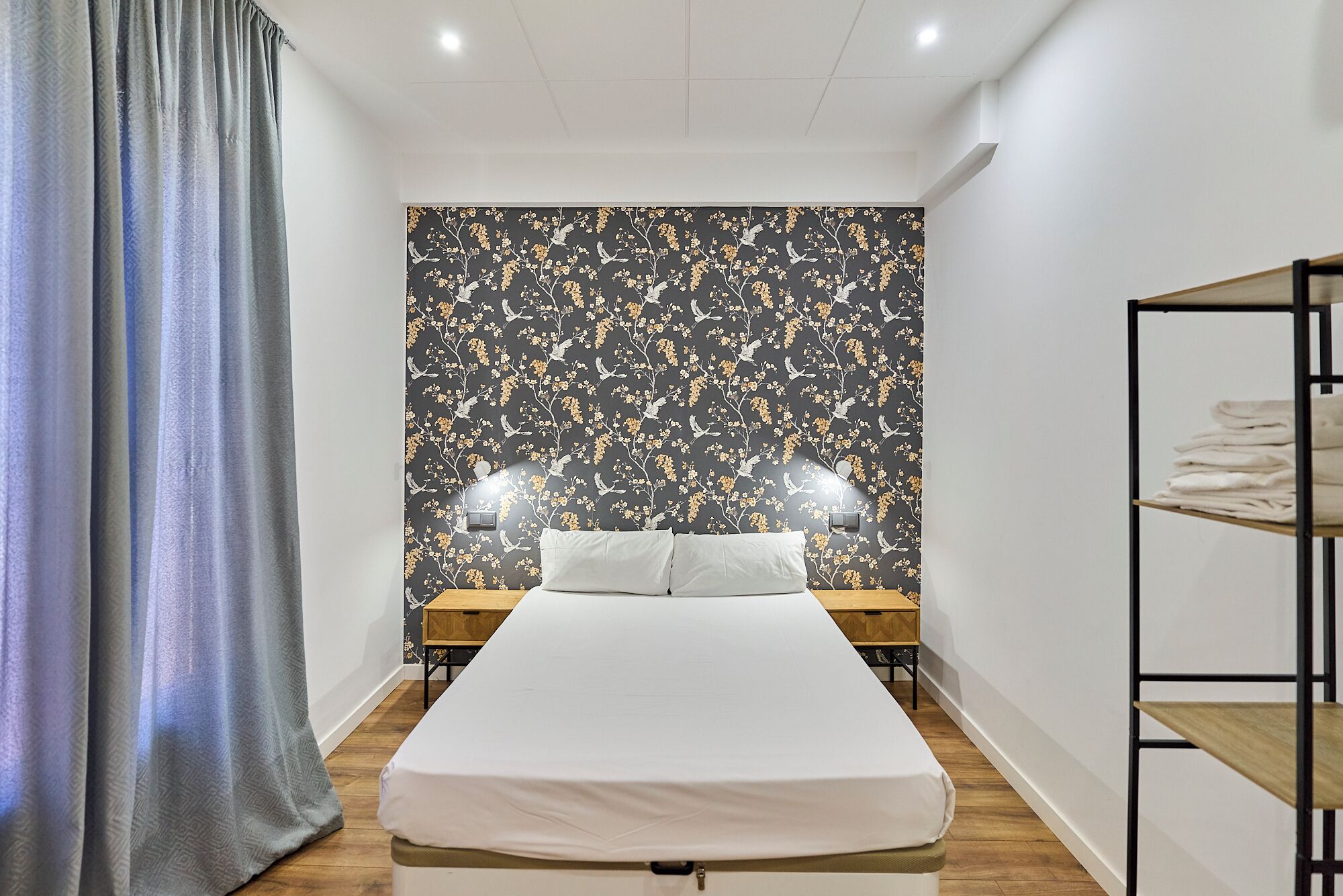 Habitación de hotel con cama, dos mesitas de noche, un estante con toallas dobladas y una ventana con cortinas.