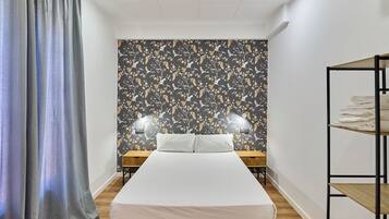 Habitación de hotel con cama, dos mesitas de noche, un estante con toallas dobladas y una ventana con cortinas.
