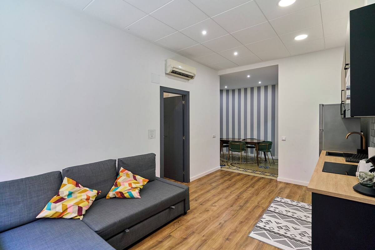 Un salon moderne avec un canapé gris, des coussins colorés, un plancher en bois, un coin cuisine avec un réfrigérateur et un mur à rayures en arrière-plan.