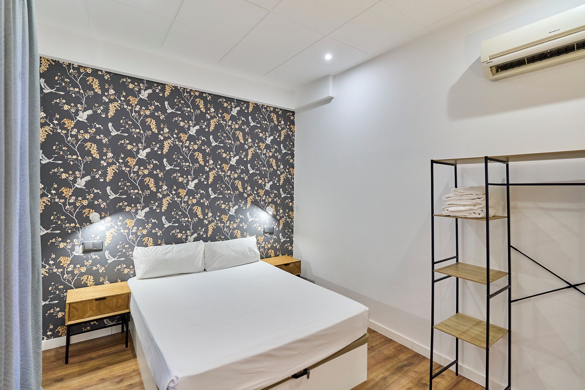 A bedroom with a bed, a nightstand, a wall-mounted shelf, and an air conditioning unit.