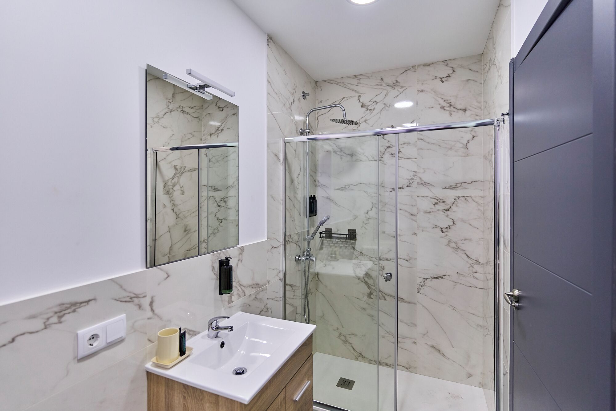 Un baño moderno con ducha envuelta en vidrio, un lavabo blanco con una taza, y un espejo.