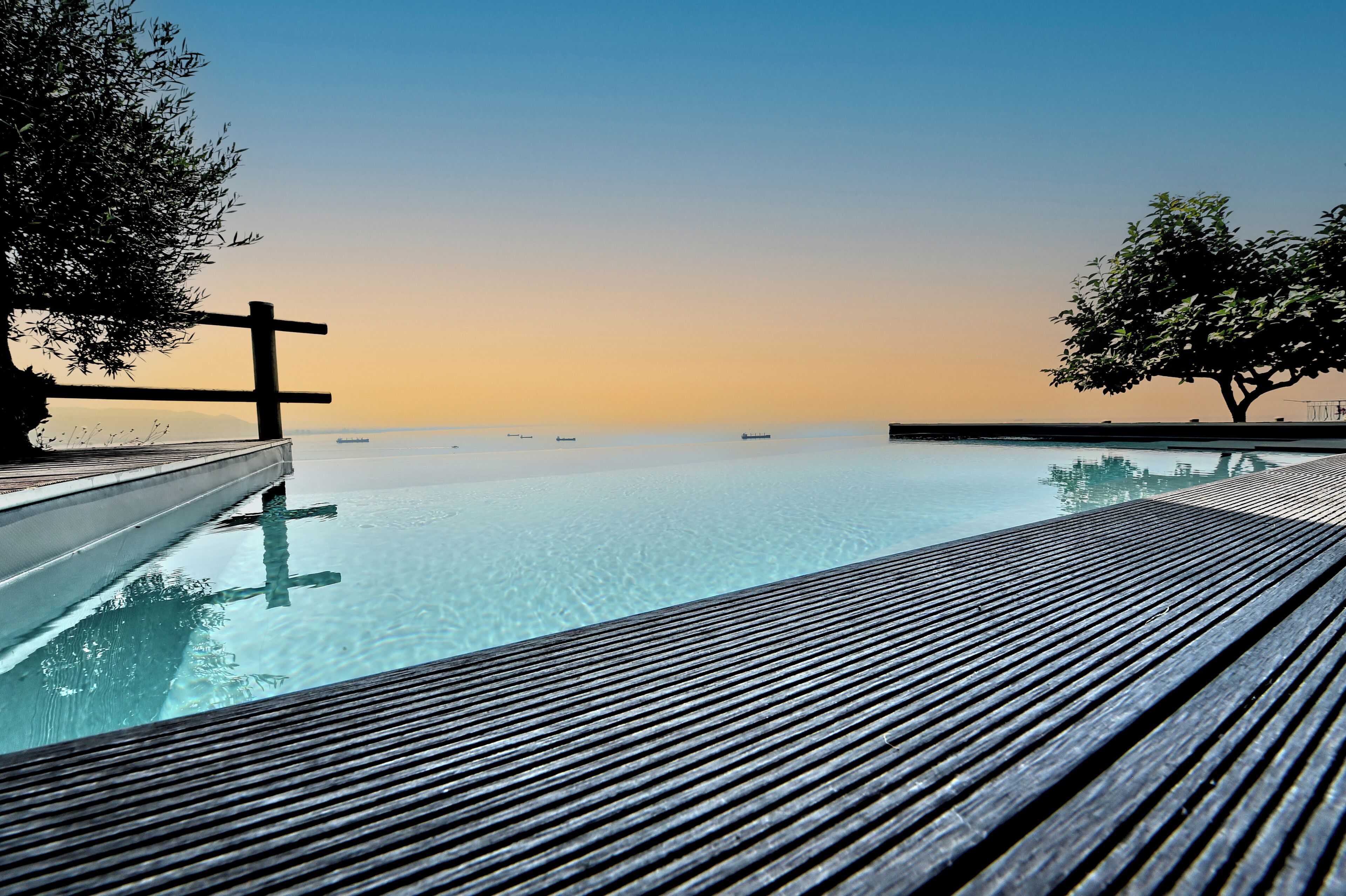 An infinity pool with a wooden deck, surrounded by trees and a clear sky.