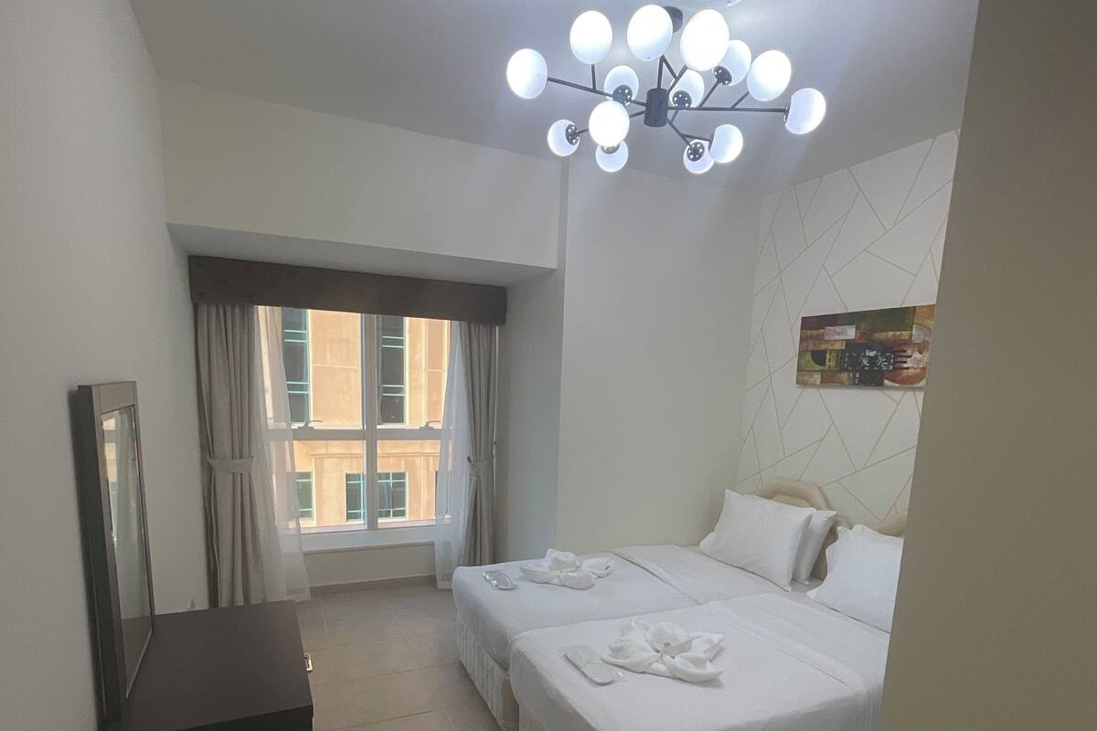 A hotel room with a bed, bedside tables, a desk, a mirror, a window with curtains, a ceiling light fixture, and a wall-mounted artwork.