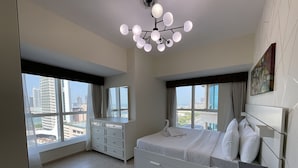 A modern bedroom with a large bed, a dresser, and a vanity. There are multiple windows providing views of the cityscape. - Marina Marvel Room Elite Residences (Dubai)