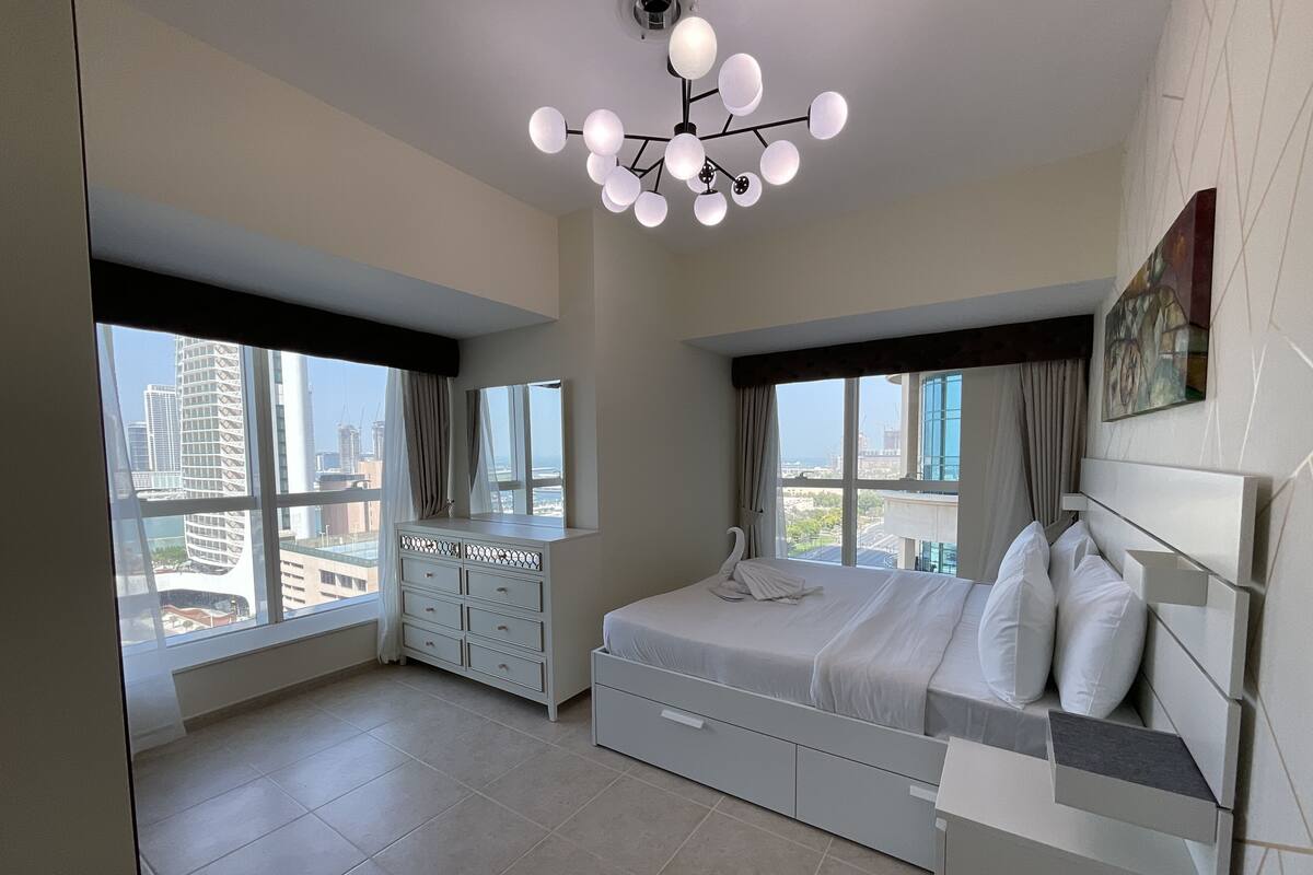 A modern bedroom with a large bed, a dresser, and a vanity. There are multiple windows providing views of the cityscape.
