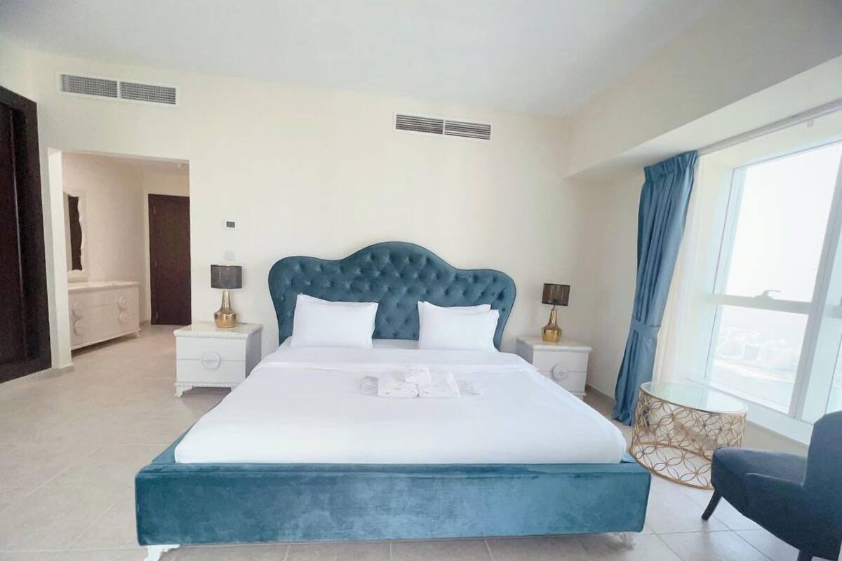 A large bed with a tufted headboard, two bedside tables with lamps, a chair, and a view of the outdoors through a window.