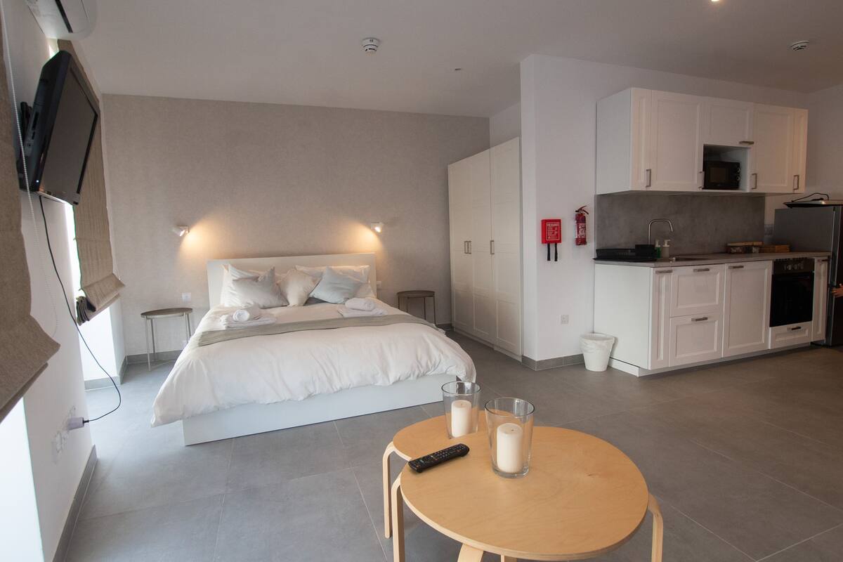 Ein modernes Hotelzimmer mit einem Bett, Nachttischen, einem runden Holztisch mit Fernbedienung und Kerzen sowie einer Küchenzeile mit weißen Schränken.