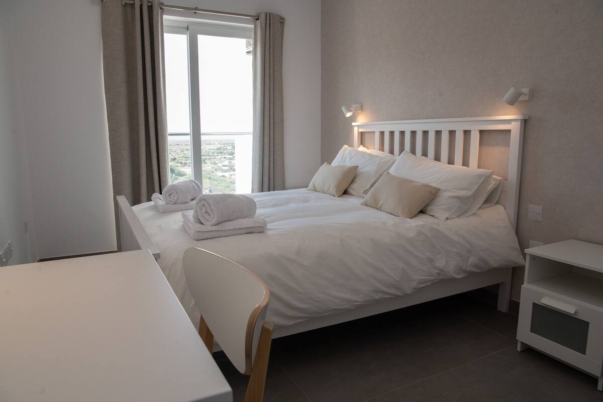 Ein ordentlich bezogenes Bett mit weißen Leinen und Kissen, ein Nachttisch, ein Stuhl und ein Beistelltischchen mit einer Lampe.